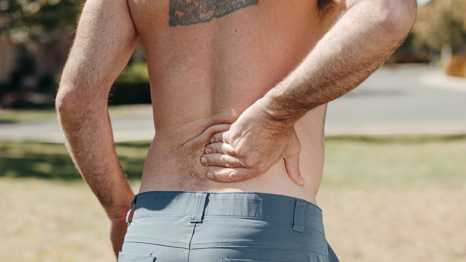 Shirtless man standing outdoors holding his lower back